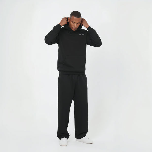 Sweat à capuche noir pour homme, modèle "Session Noire" de la marque Tourismo Clothing. Hoodie sans cordon, coupe droite, capuche structurée et poche kangourou. Logo brodé en blanc "TOURISMO" avec effet damier placé sur la poitrine gauche. Le mannequin ajuste sa capuche, tête baissée, bras relevés. Ensemble porté avec un pantalon noir ample et des sneakers blanches. Photo prise en studio sur fond blanc, lumière neutre. Tenue streetwear sobre, inspirée des ambiances de circuits fermés et des sessions de test